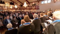 Een kerk vol met mensen en een koor met blauw sjaaltjes en kroonluchters aan het plafond