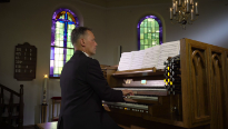 Man in pak in een niet verlicht kerk spelend op een orgel met drie lagen toetsen en registers en een kroonluchter in de kerk